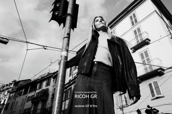 Portrait en noir et blanc d'une jeune femme en contre-plongée, posant dans un environnement urbain, avec la mention "Camera powered by RICOH GR" et "realme GT 8 Pro"