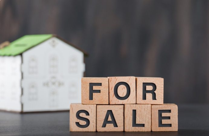 Cubes en bois formant les mots For Sale devant une maison miniature, illustrant la corrélation entre taxe fonciere et baisse valeur bien.